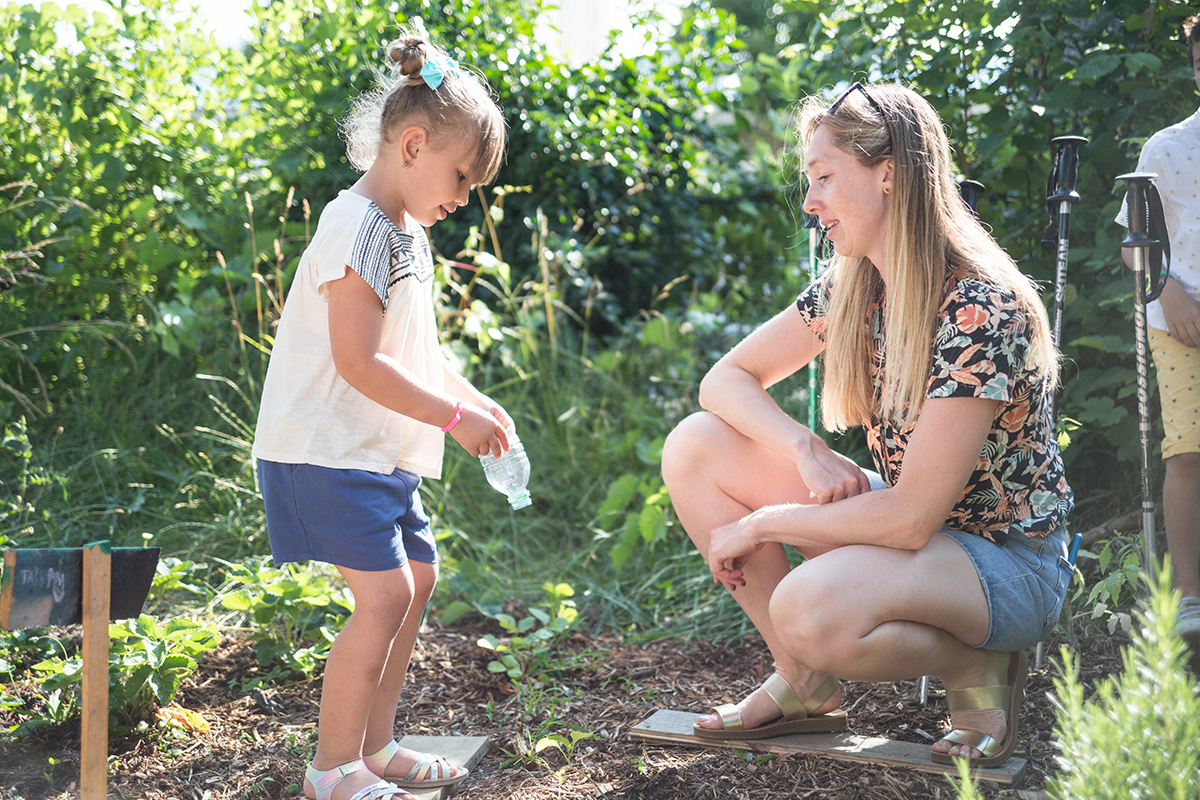 Une femme jardine avec une jeune enfant qui arrose des plantes dans un potager